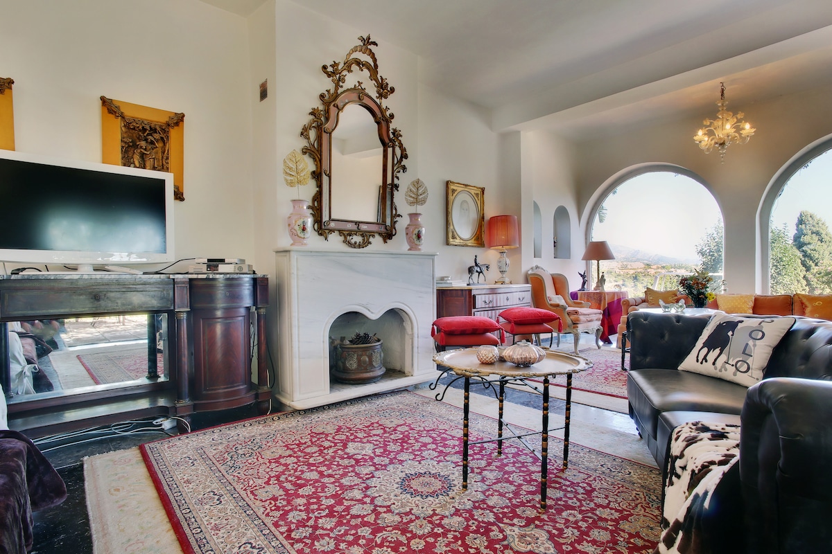 A cozy living room presents a classic fireplace framed by an ornate mirror. Plush seating options, including a black leather couch and vibrant red chairs, invite relaxation. Large arched windows offer views of the surrounding landscape, while an elegant chandelier provides warm light overhead.