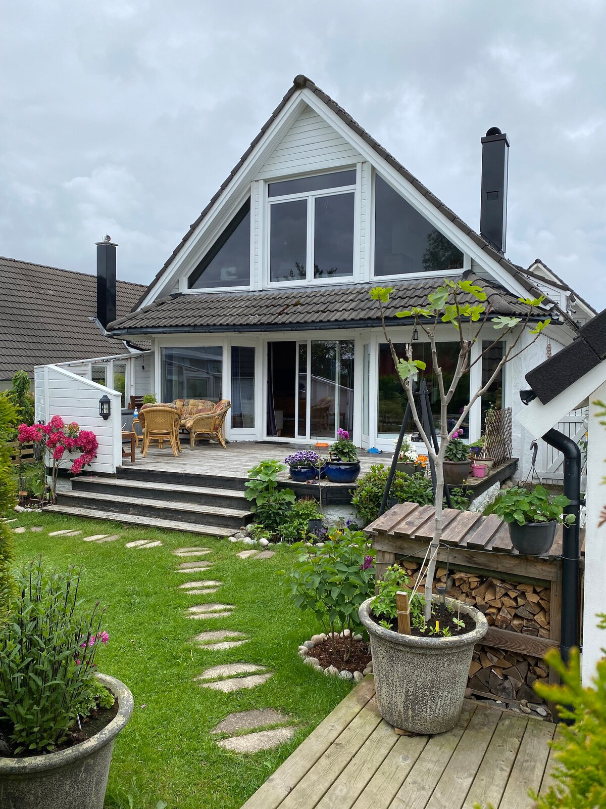 The exterior of a large house features a sloped roof and large glass windows that offer a view of the garden. Stone steps lead to a porch area with wooden seating surrounded by potted plants and flowers. A well-maintained lawn and garden area are visible.