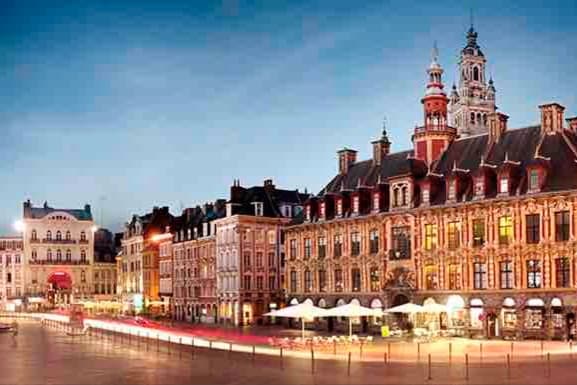 A vibrant square is featured, showcasing historical buildings with intricate architectural details. Warm lights illuminate the façades, and tables with umbrellas are set outside. The sky transitions to twilight, providing a soft backdrop to the lively atmosphere of the area.