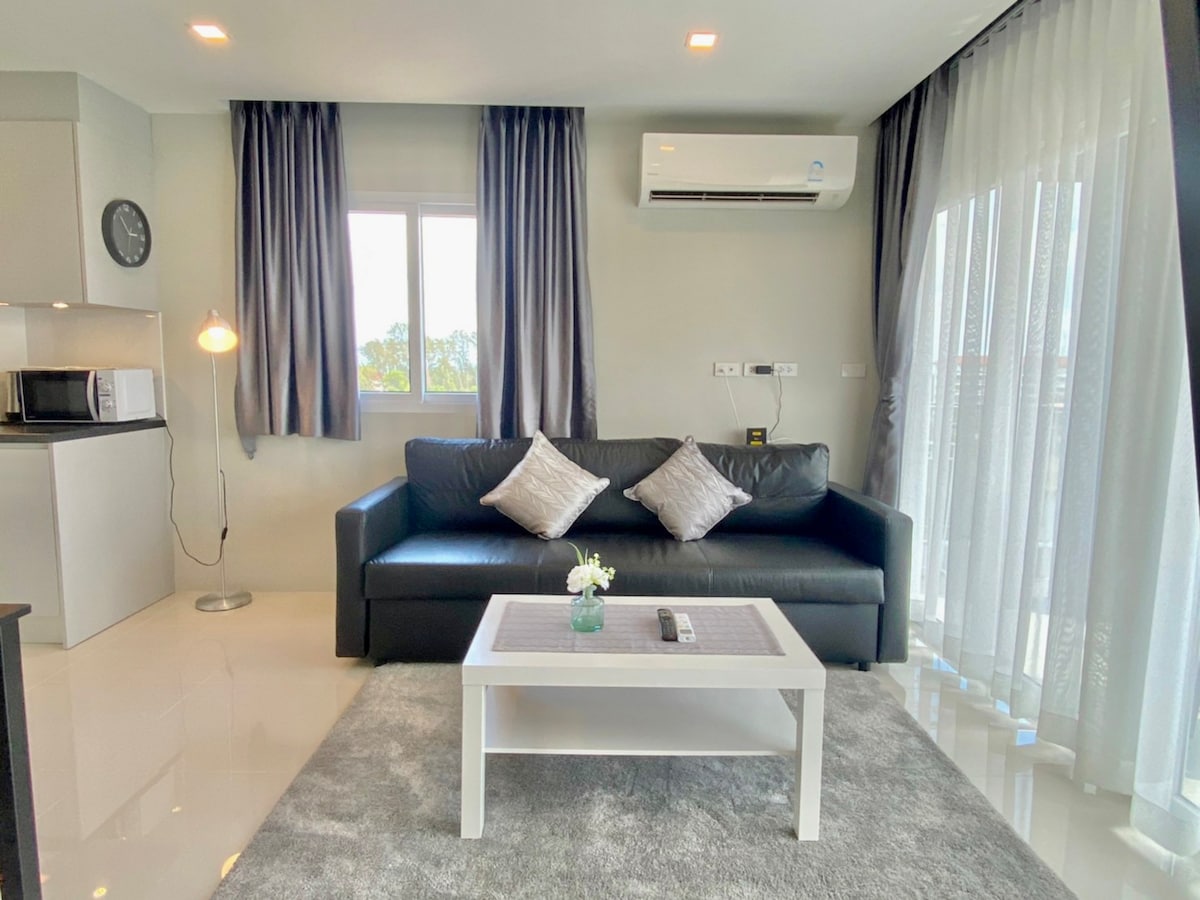 A modern living area is seen featuring a sleek black sofa with decorative cushions. A white coffee table is positioned in front, and a small flower vase adds a touch of charm. Natural light streams through sheer curtains, highlighting the gleaming tiled floor.
