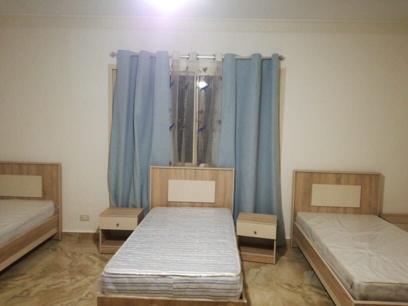 A bright room is furnished with three beds, two of which have mattresses. Light blue curtains frame a window that allows natural light to fill the space. Two nightstands are positioned beside the beds, enhancing the room's functionality.