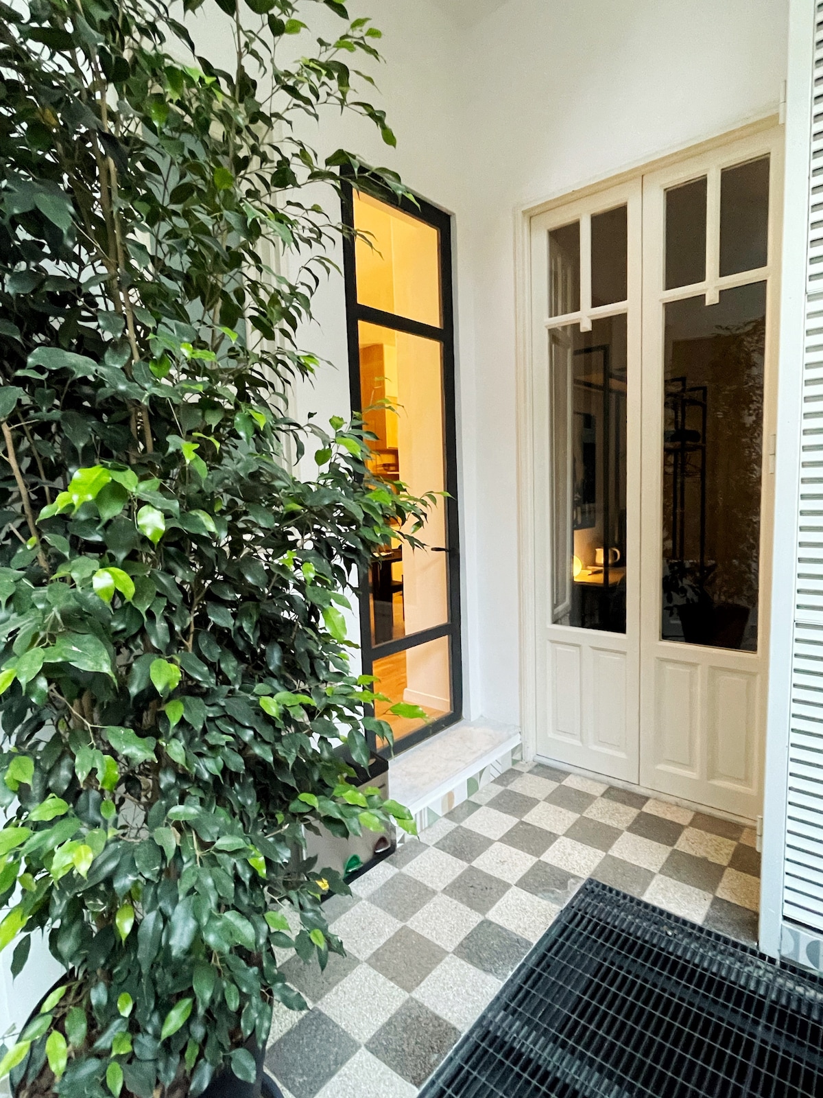 The entrance features a checkerboard tile floor leading to a double door framed by large leafy plants. Soft lighting from inside creates a welcoming ambiance, highlighting the elegant design of the door frames and the warm colors of the interior.