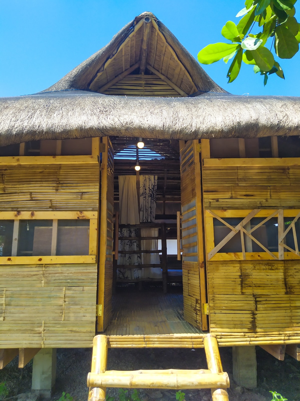 A beach hut is shown with a thatched roof, showcasing wooden walls and a wooden walkway. Entry is framed by open doors, allowing natural light to illuminate the interior. The structure is set against a clear blue sky with hints of green foliage in the foreground.