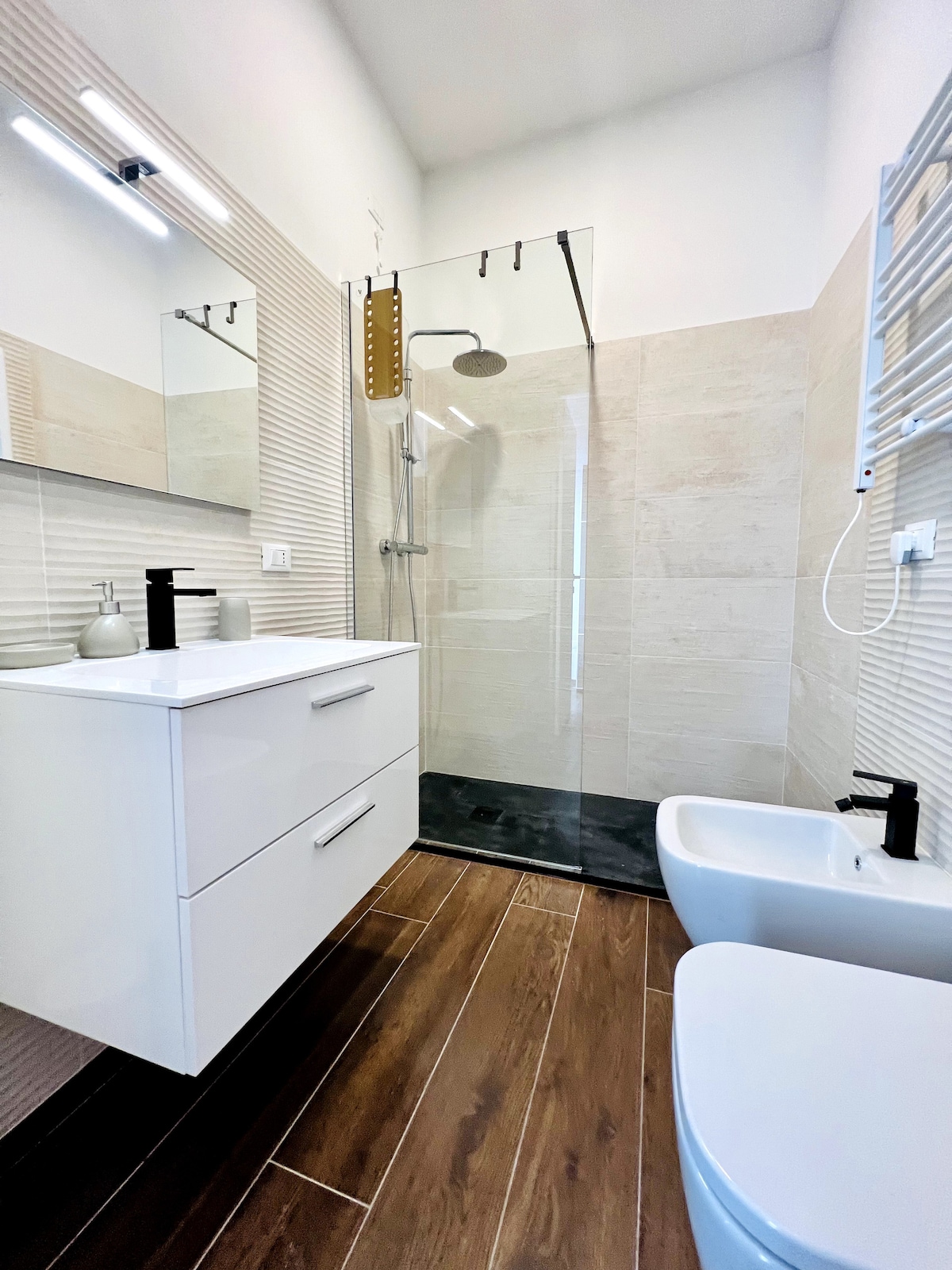 A clean and modern bathroom features a walk-in shower with a glass partition. A vanity with a sleek countertop is complemented by a large mirror. Dark wood flooring contrasts with light-colored tiles, creating a spacious and bright environment. Amenities include a bidet and contemporary fixtures.