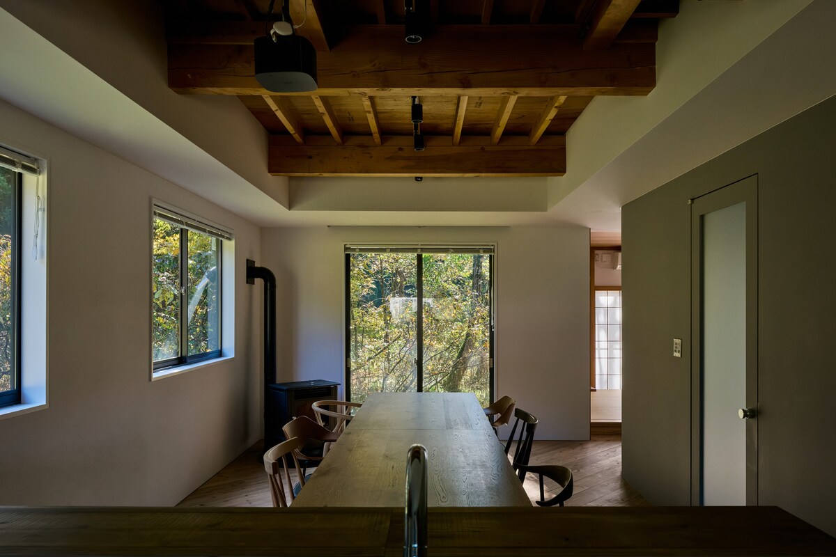 A spacious dining area features a long wooden table surrounded by several chairs. Large windows allow natural light to fill the room, offering views of the greenery outside. Exposed wooden beams add a rustic element to the contemporary design.