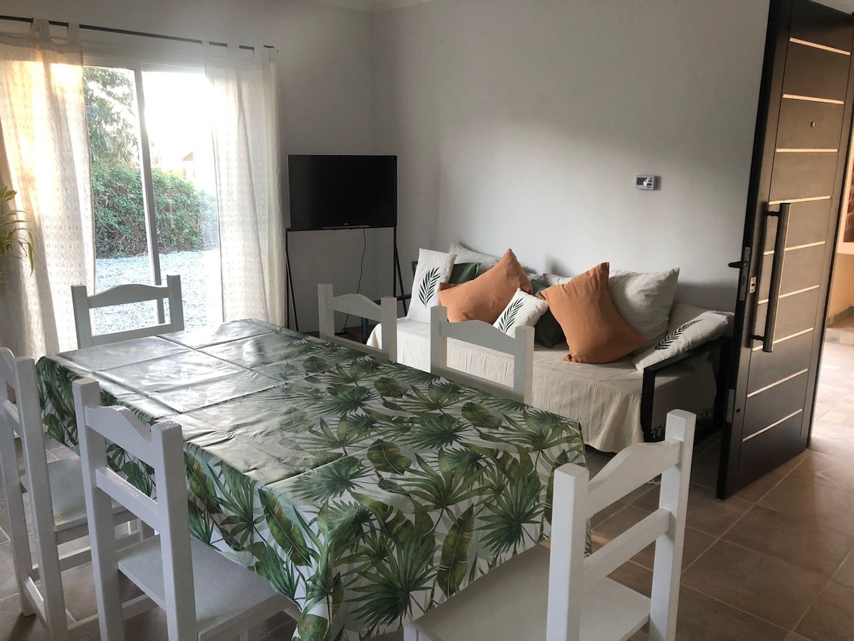 An open living space features a dining table with a tropical-themed tablecloth, set for multiple diners. A light-filled area includes a sofa adorned with decorative cushions and a television mounted on the wall. Large windows offer views of the surrounding greenery.