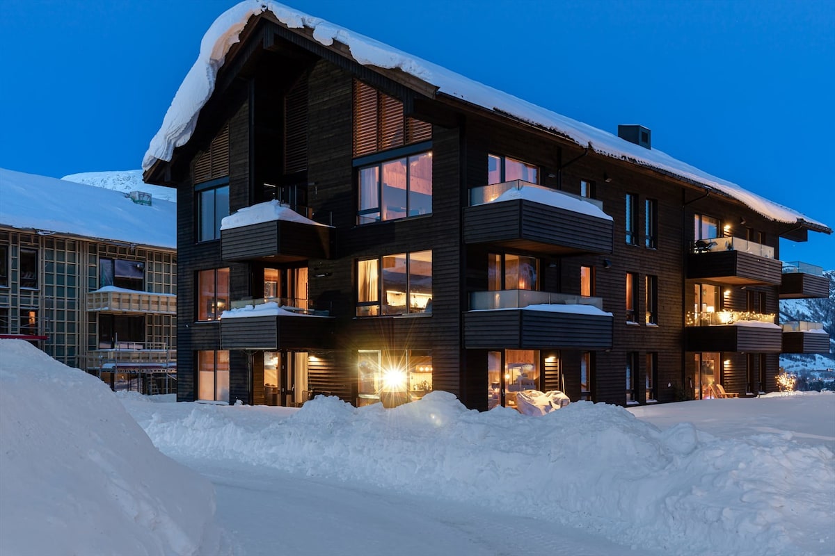 A modern chalet-style building is set against a snowy landscape, showcasing multiple balconies with glass railings. Warm lights illuminate the interiors through large windows, creating a welcoming contrast to the cold exterior. The structure has a sloped roof, characteristic of alpine architecture.
