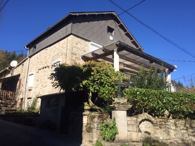 A stone house is seen from the side, featuring a covered terrace and a well-maintained garden area. The structure is characterized by a pitched roof and several windows, blending harmoniously with the surrounding green landscape under a clear blue sky.