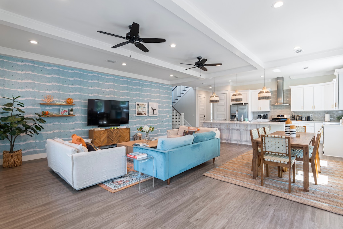 An airy living area features two seating areas, one with a blue sofa and the other with a white sofa, both arranged around a central area rug. A dining table sits nearby, complemented by pendant lighting. Light blue walls and modern decor enhance the open space.