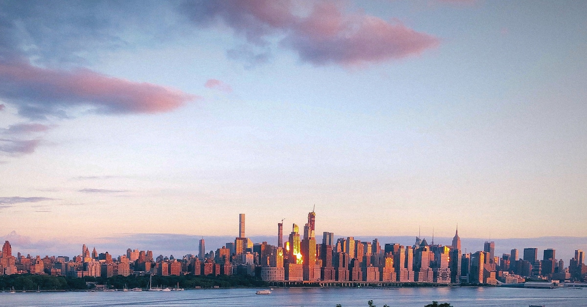 A panoramic view of the Manhattan skyline is captured, showing tall buildings and the Hudson River in the foreground. The scene is illuminated by the soft glow of the sunset, highlighting the architectural silhouettes against a colorful sky.