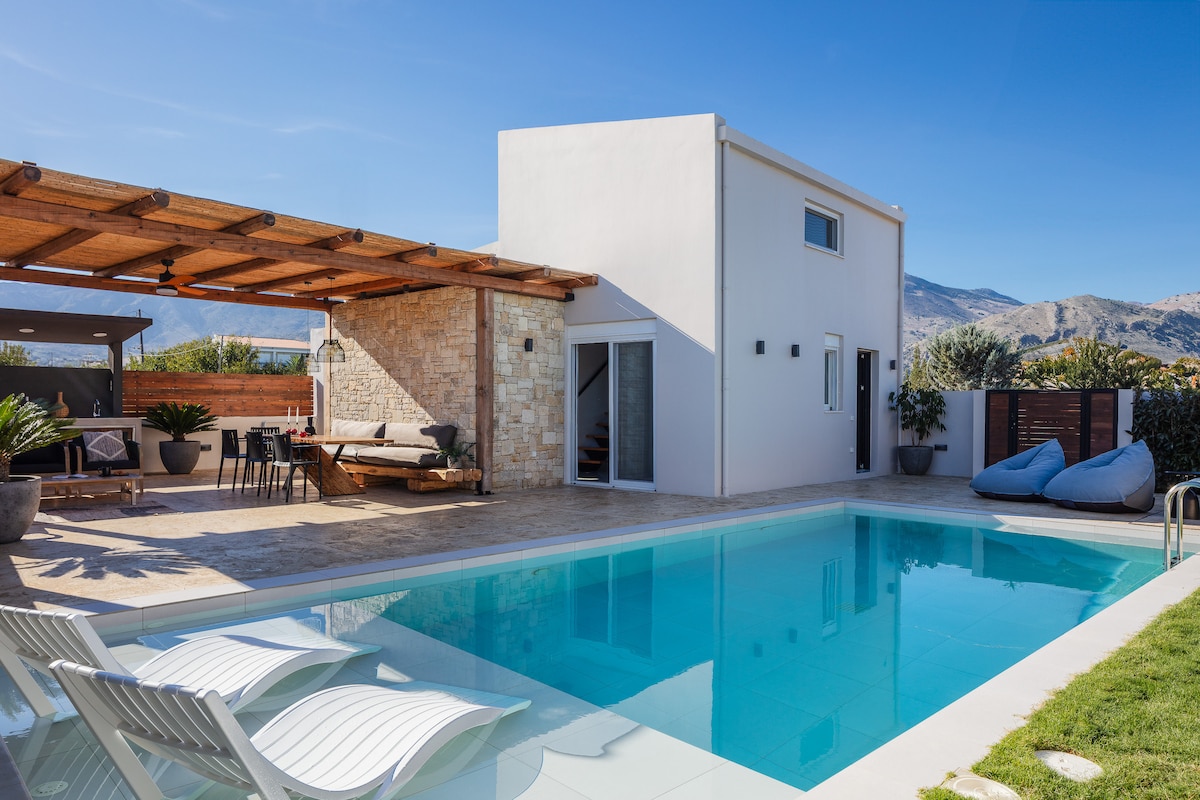 The outdoor area features a private swimming pool surrounded by lounge chairs, offering a space for relaxation. A covered terrace provides shade, with a dining table and seating, complemented by natural stone and greenery. The mountains are visible in the background against a clear blue sky.