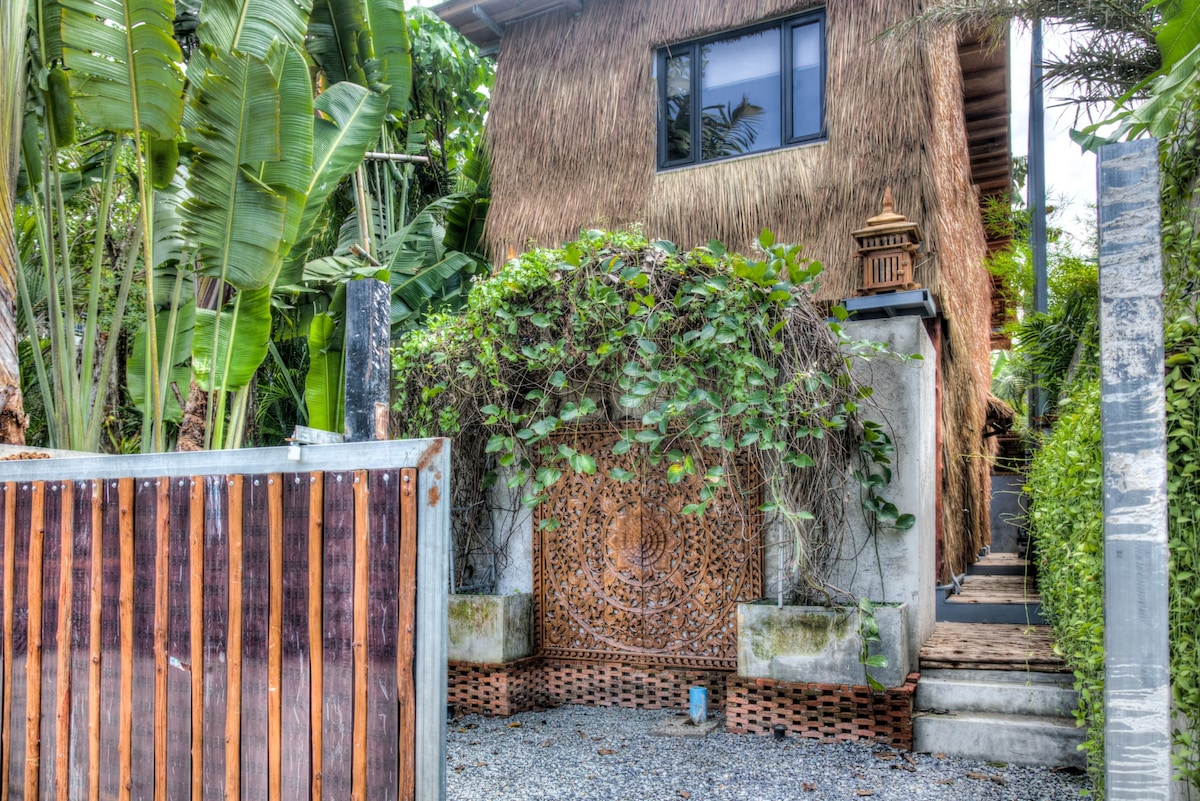 The inviting entrance features a carved stone wall surrounded by lush greenery. A rustic wooden gate provides access to the pathway leading to the bungalow. Tall banana plants frame the area, enhancing the natural atmosphere of the property.