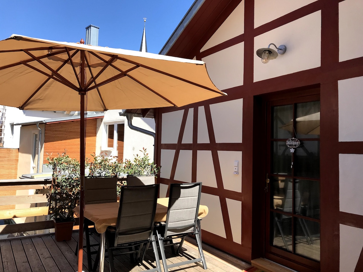 A spacious wooden terrace is showcased, featuring a large umbrella over a dining table with several chairs. The terrace is adorned with plants and framed by traditional half-timbered architecture, providing a blend of outdoor comfort and historic charm.
