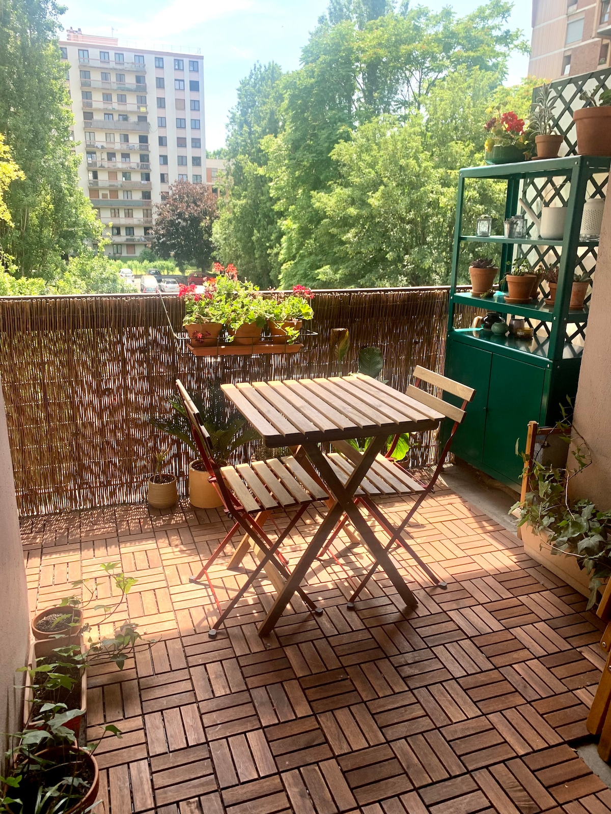 A balcony area is presented with a small wooden table and two chairs, surrounded by potted plants and flowers. The flooring consists of wooden tiles, and greenery from nearby trees adds a serene backdrop.