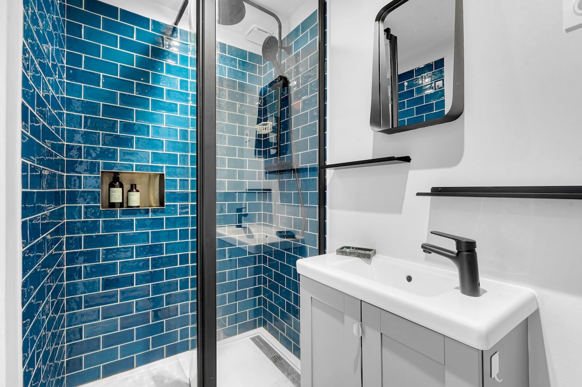 A modern bathroom features a walk-in shower with striking blue tiled walls. A sleek black mirror and minimalist sink are mounted against a contrasting white backdrop. An inset shelf holds essential toiletries, adding functionality to the stylish space.