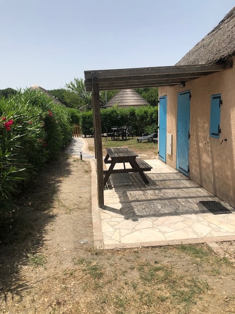 A pathway leads to a charming outdoor seating area featuring a shaded picnic table under a wooden pergola. Lush greenery surrounds the space, providing privacy. The exterior walls of the building are painted in a soft hue, complemented by vibrant blue doors.