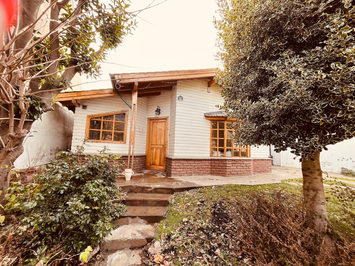 An inviting exterior of a single-story house is displayed, featuring a wooden door framed by large windows. A well-maintained garden with greenery surrounds the entrance, and stone steps lead up to the porch, providing a warm and welcoming atmosphere.