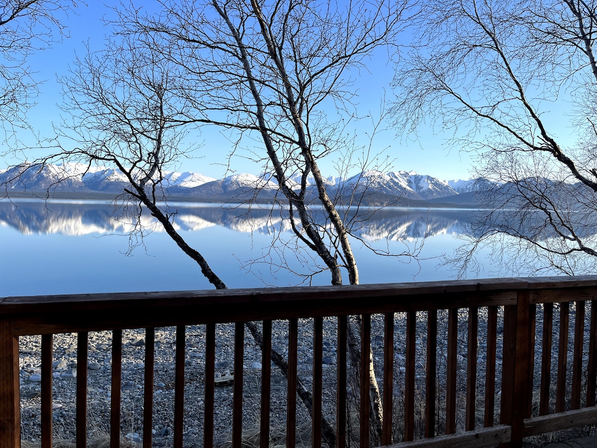 A serene view of a calm lake reflects surrounding snow-capped mountains under a clear blue sky. Leafless trees frame the foreground, while a wooden railing adds a natural touch to the picturesque landscape.