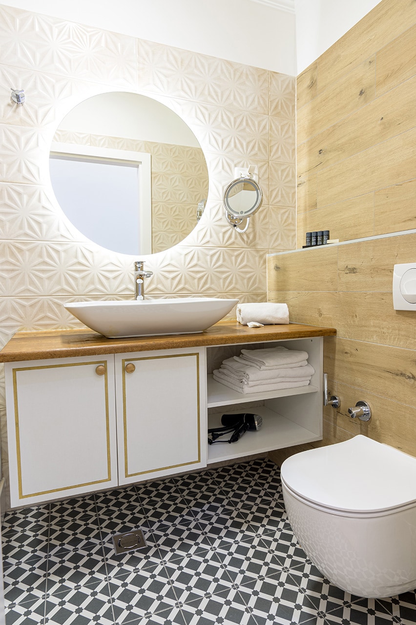 A modern bathroom is showcased with a stylish circular mirror above a basin. Wooden accents contrast with geometric patterned tiles on the floor. Fresh towels are neatly arranged on a shelf, and toiletries are provided for convenience.