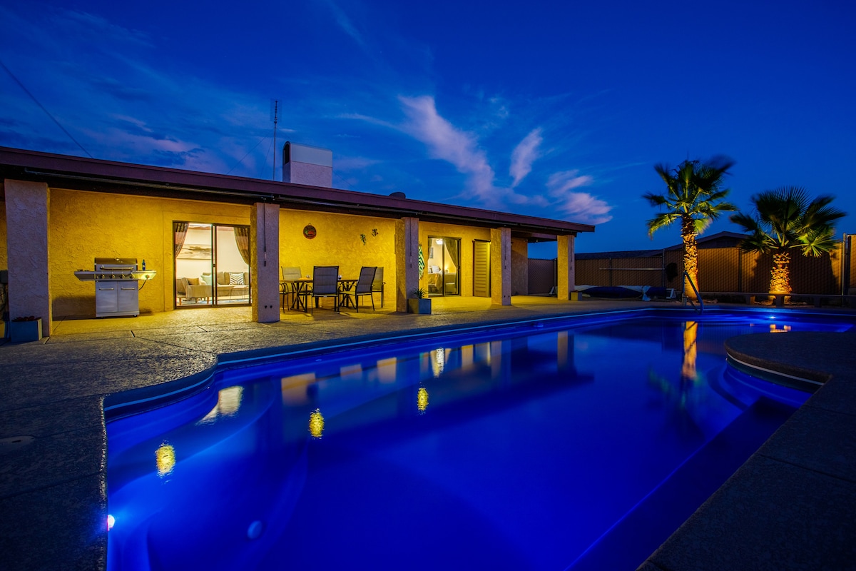An inviting outdoor area is showcased at dusk, featuring a shimmering pool illuminated by soft blue lighting. The patio area includes a dining set and a barbecue grill, with palm trees gently framing the space, complementing the tranquil atmosphere.