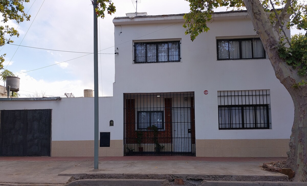 The exterior of the house is presented, showcasing a two-story white structure with a gated entrance. Large windows with black grilles are featured, allowing natural light into the interior. A tree shade is visible alongside the walkway, providing a sense of greenery.