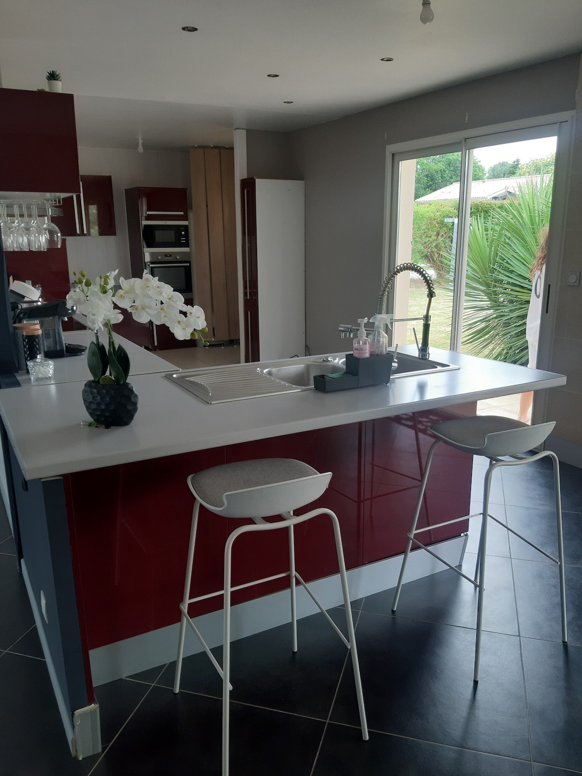 A modern kitchen features sleek red cabinetry and a central island with seating for two. A sliding glass door allows natural light to enter and provides a view of the outdoor greenery. Elegant white orchids are displayed on the counter.