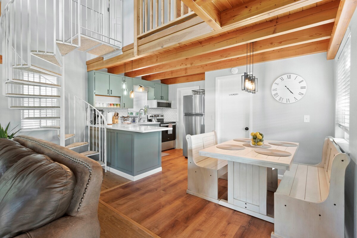 An open kitchen and dining area is featured with light wood beams overhead. A spiral staircase leads to the loft space above. The dining table, designed with built-in benches, is set for meals, while modern appliances and sleek cabinetry are visible in the kitchen area.