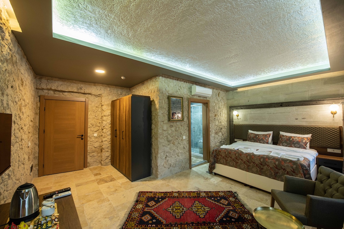 A spacious hotel room features stone walls and a large bed dressed in elegant linens. A sitting area includes a plush armchair beside a decorative rug. The room is illuminated by soft ceiling lights, and a wooden closet is positioned near the entrance.
