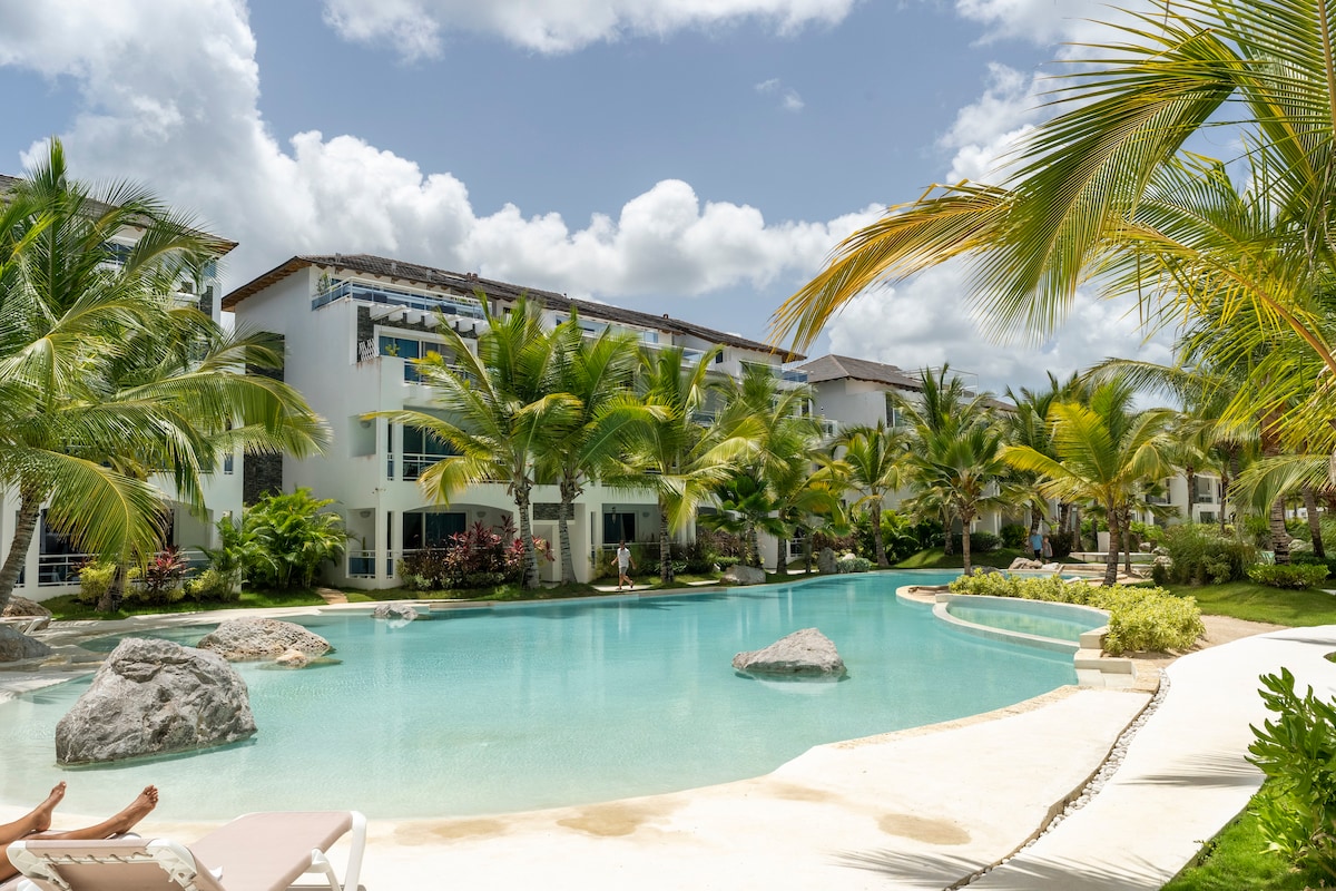 A tranquil pool area is surrounded by lush palm trees and colorful vegetation, with large rocks scattered throughout the water. The building features multiple balconies overlooking the serene landscape, providing a peaceful setting for relaxation.