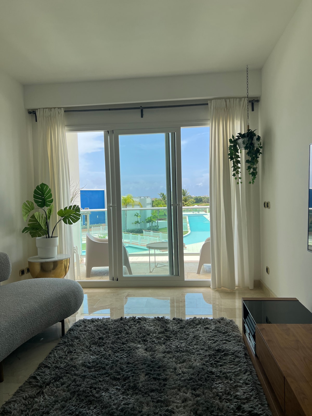 A bright living area features large sliding glass doors opening to a swimming pool view. Light curtains frame the entrance, while a plush rug adds texture to the floor. A sofa and potted plant contribute to the room's modern design.