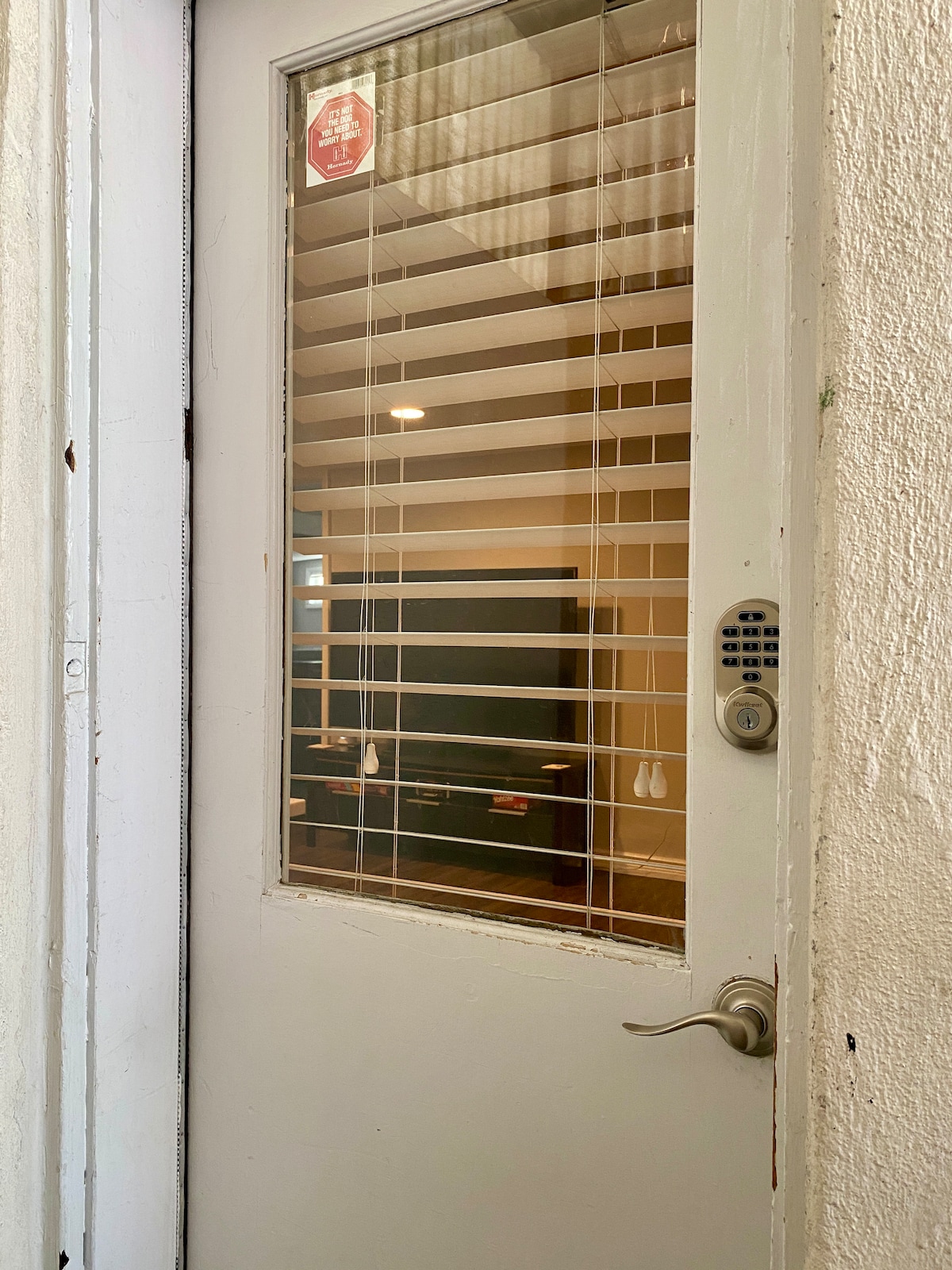 The image depicts a white door with a glass panel featuring horizontal blinds. A keypad entry system is visible alongside the door handle. The interior is faintly illuminated, suggesting a welcoming atmosphere within the apartment.