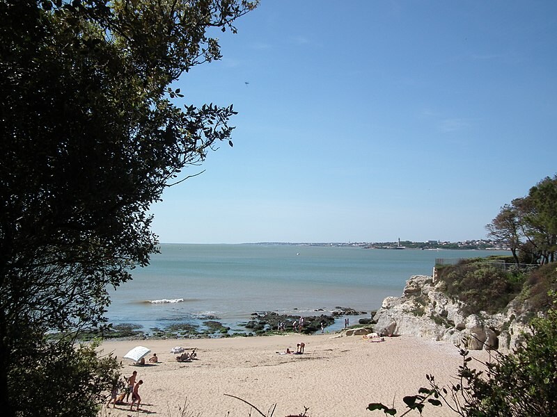 A sandy beach is visible beyond lush greenery, with gentle waves lapping at the shore. People are scattered across the beach, relaxing under the sun. The tranquil sea extends into the distance, revealing a hint of coastal scenery beyond.