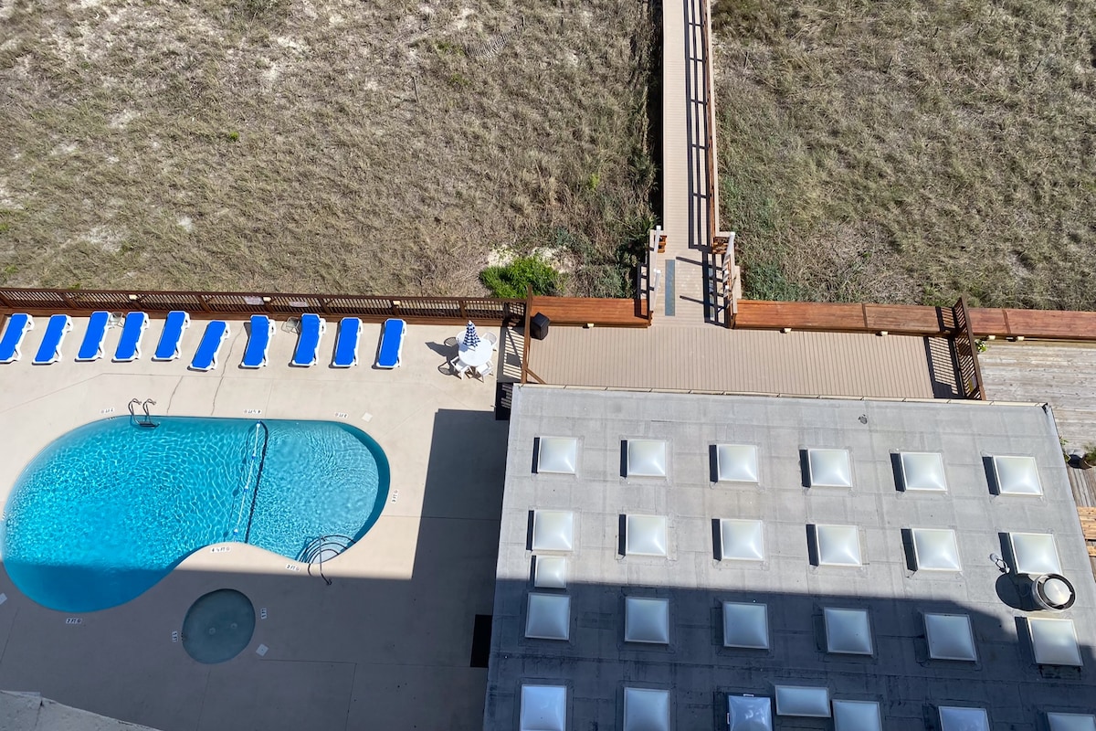 An aerial view captures a curved swimming pool surrounded by multiple lounge chairs arranged for comfort. A wooden walkway leads to a sandy area, with a portion of a large, flat-roofed structure visible, featuring a series of shaded seating areas.