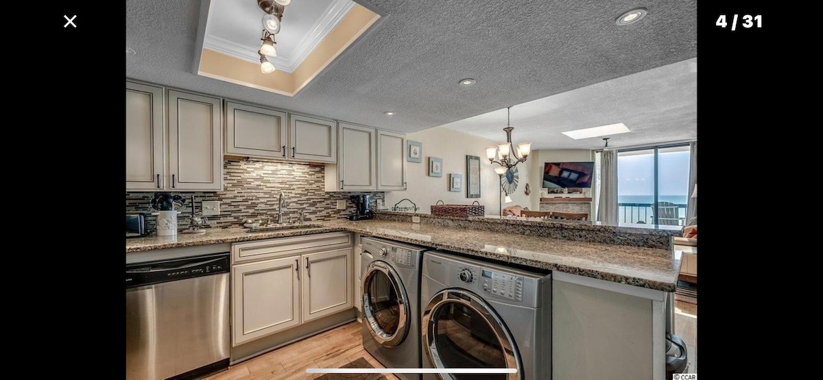 A contemporary kitchen space is equipped with stainless steel appliances, including a dishwasher and front-loading washer and dryer. Light-colored cabinetry contrasts with the granite countertops. Natural light filters through the nearby windows, highlighting the open layout that connects to a dining and living area.