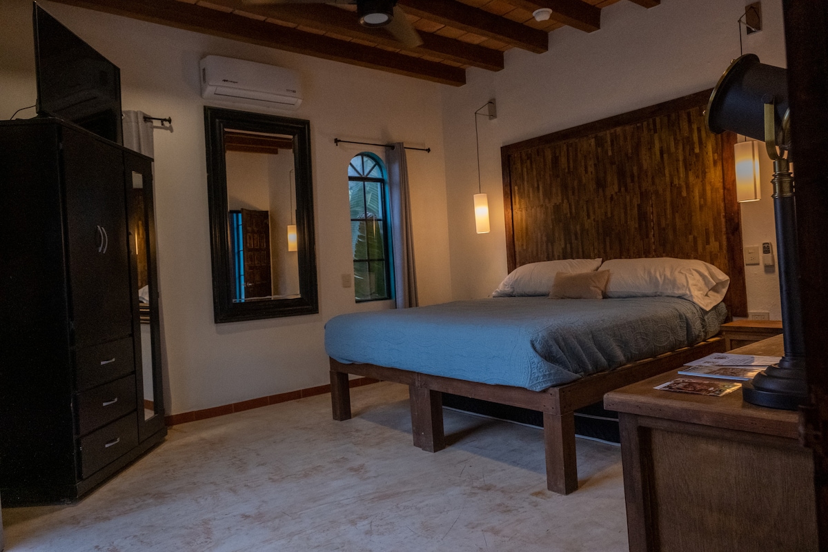 A spacious King Size bed is positioned against a textured wooden headboard. A large mirror is mounted on the wall, reflecting natural light. A wardrobe and a bedside lamp are visible, contributing to the room's functional layout. Soft tones create a calm environment.