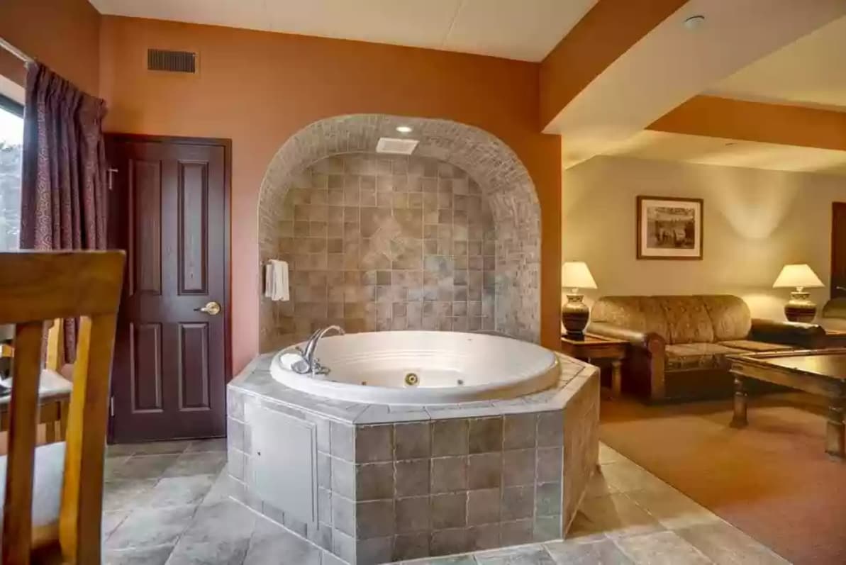 A spacious corner of the living area features a large, round jacuzzi set within a stone alcove. Soft lighting complements the warm tones of the walls, while a cozy seating arrangement with a sofa is visible in the background. A window allows natural light to enhance the space.