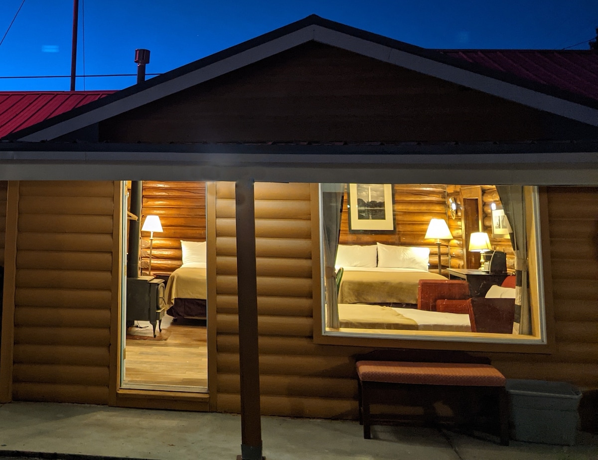 The exterior of a log cabin showcases a warm glow from interior lighting. Two large windows reveal a cozy interior with two queen beds, nightstands, and chair seating. A bench is positioned outside, and the structure features a welcoming roofline against a twilight sky.