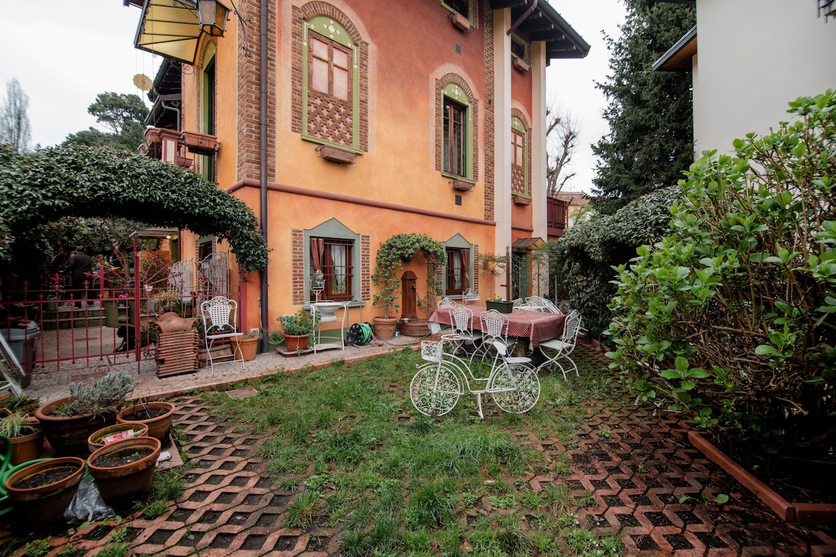 A tranquil garden area is visible, featuring a variety of potted plants and a manicured lawn. Rustic furniture, including a table and chairs, can be seen under shaded areas. The picturesque facade of the house, adorned with decorative windows, completes the peaceful setting.