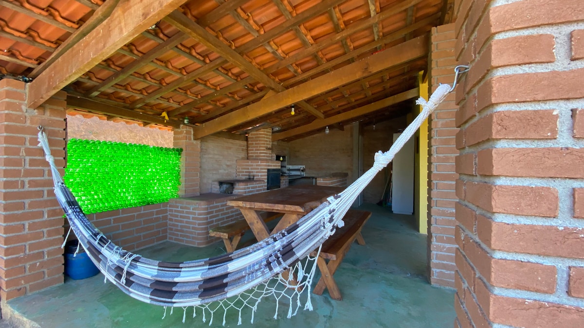 An open outdoor space features a wooden dining table surrounded by benches, with a braided hammock hanging nearby. A stone grill is positioned in the background, and a vibrant green wall composed of bottles adds a unique decorative touch.