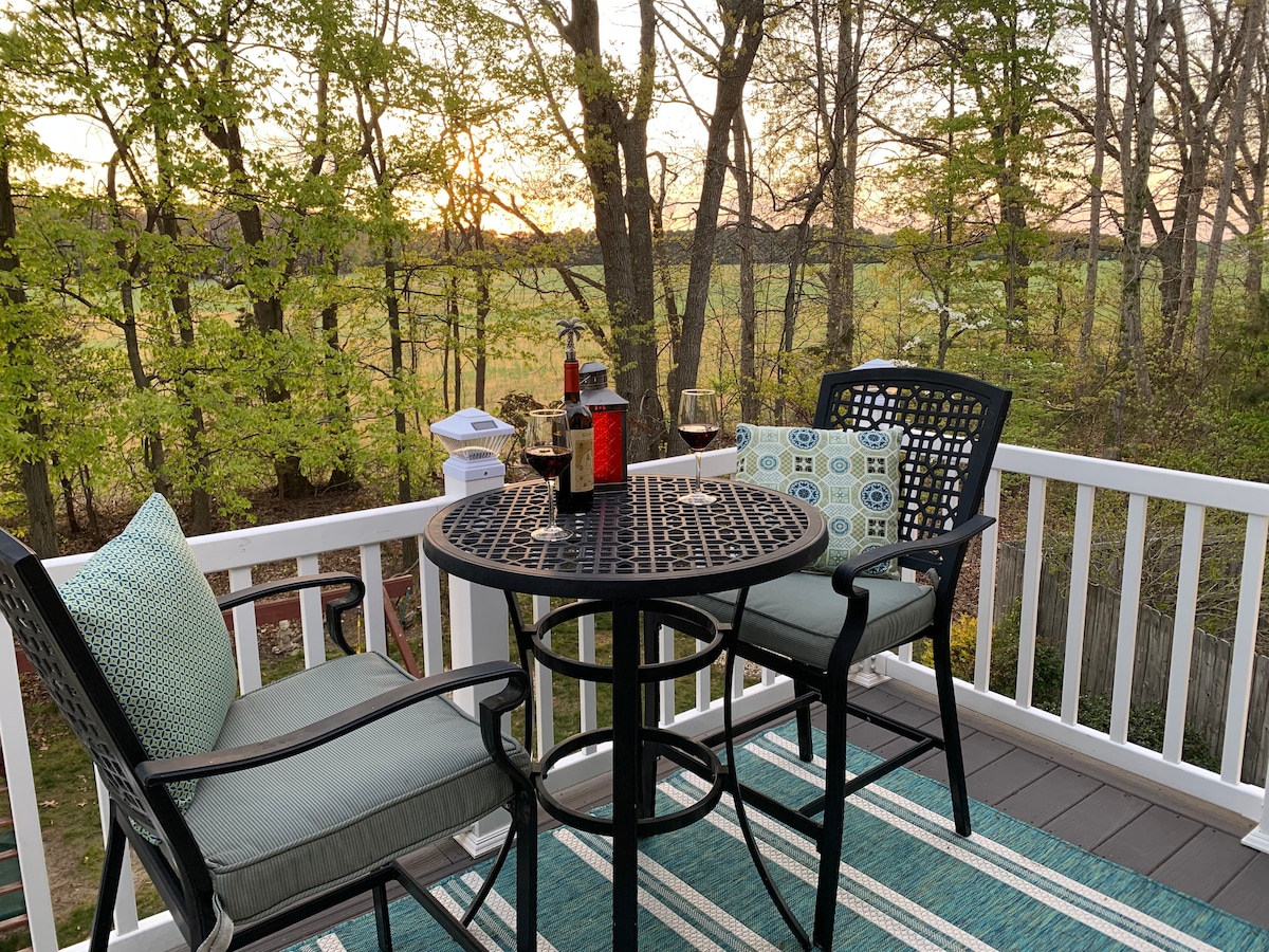 A charming deck is furnished with a small round table and two comfortable chairs, surrounded by lush greenery. A bottle and glasses are placed on the table, offering a scenic viewpoint of the sunset through the trees.
