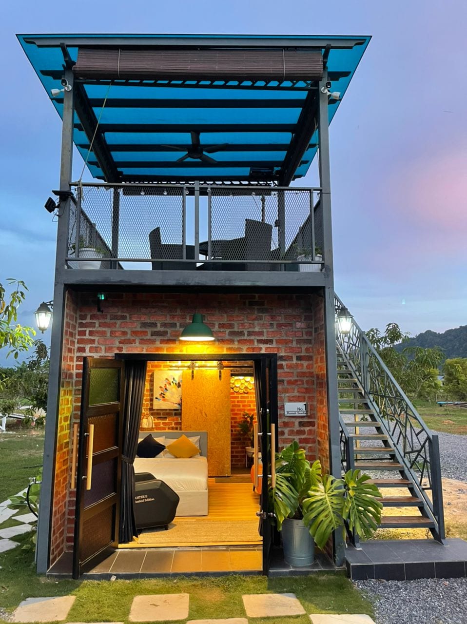 A modern two-story cabin features a distinctive brick façade with a welcoming entrance. Stairs lead up to a rooftop terrace, furnished with seating. Large windows provide natural light to the interior area, showcasing a comfortable sleeping space decorated with neutral tones.