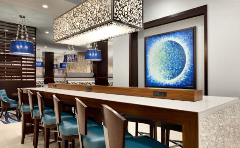 An elegant dining area features a long white stone countertop with ample seating. Stylish blue chairs complement the intricate patterned pendant lights above. A vibrant abstract artwork in shades of blue and green adorns the wall, adding a contemporary touch to the space.