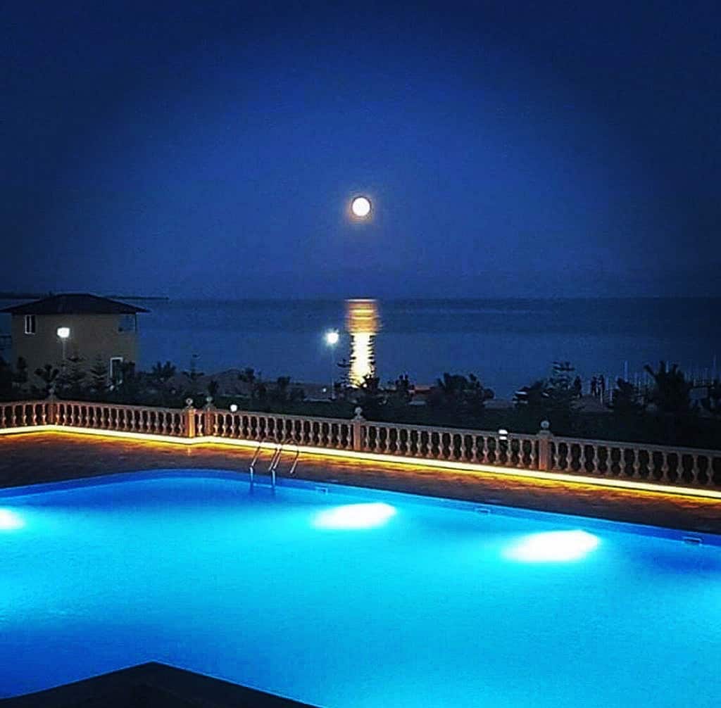 A serene evening view features a glowing full moon reflecting on the calm lake surface. The tranquil scene is complemented by illuminated areas around a swimming pool, casting a soft blue light in the night.