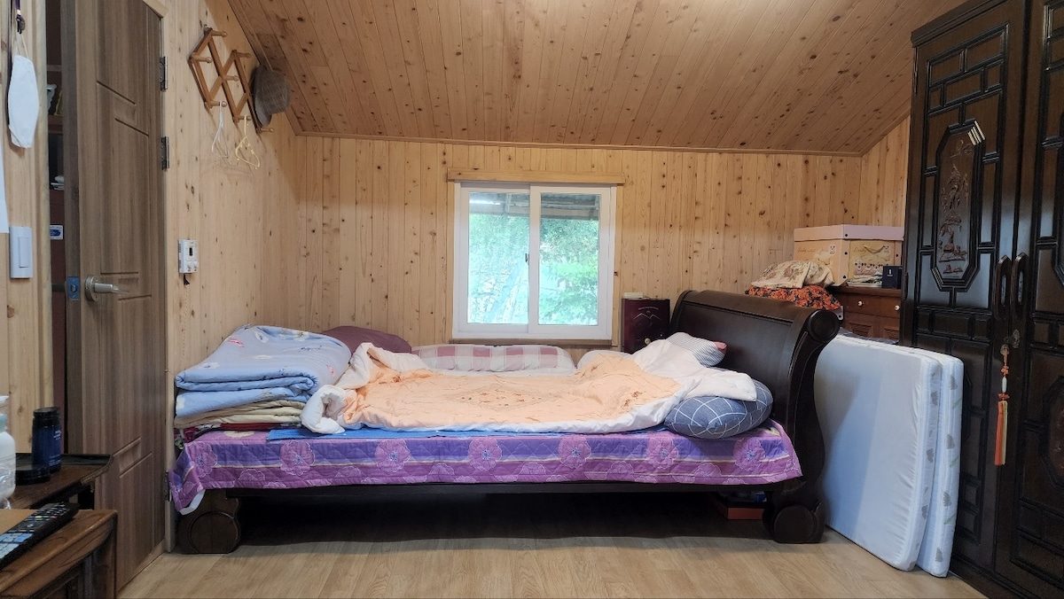 A spacious sleeping area features a large bed with layered bedding and pillows. Natural wood paneling lines the walls, while a window allows soft light to enter the room. Nearby furniture includes a wooden cabinet and a dresser, contributing to the cozy atmosphere.