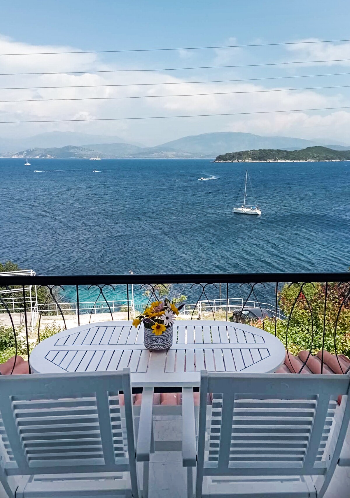 A balcony features a round white table and two chairs, with a vase of fresh flowers at the center. The expansive ocean view showcases sailboats on a clear blue sea, with distant islands under a partially cloudy sky.
