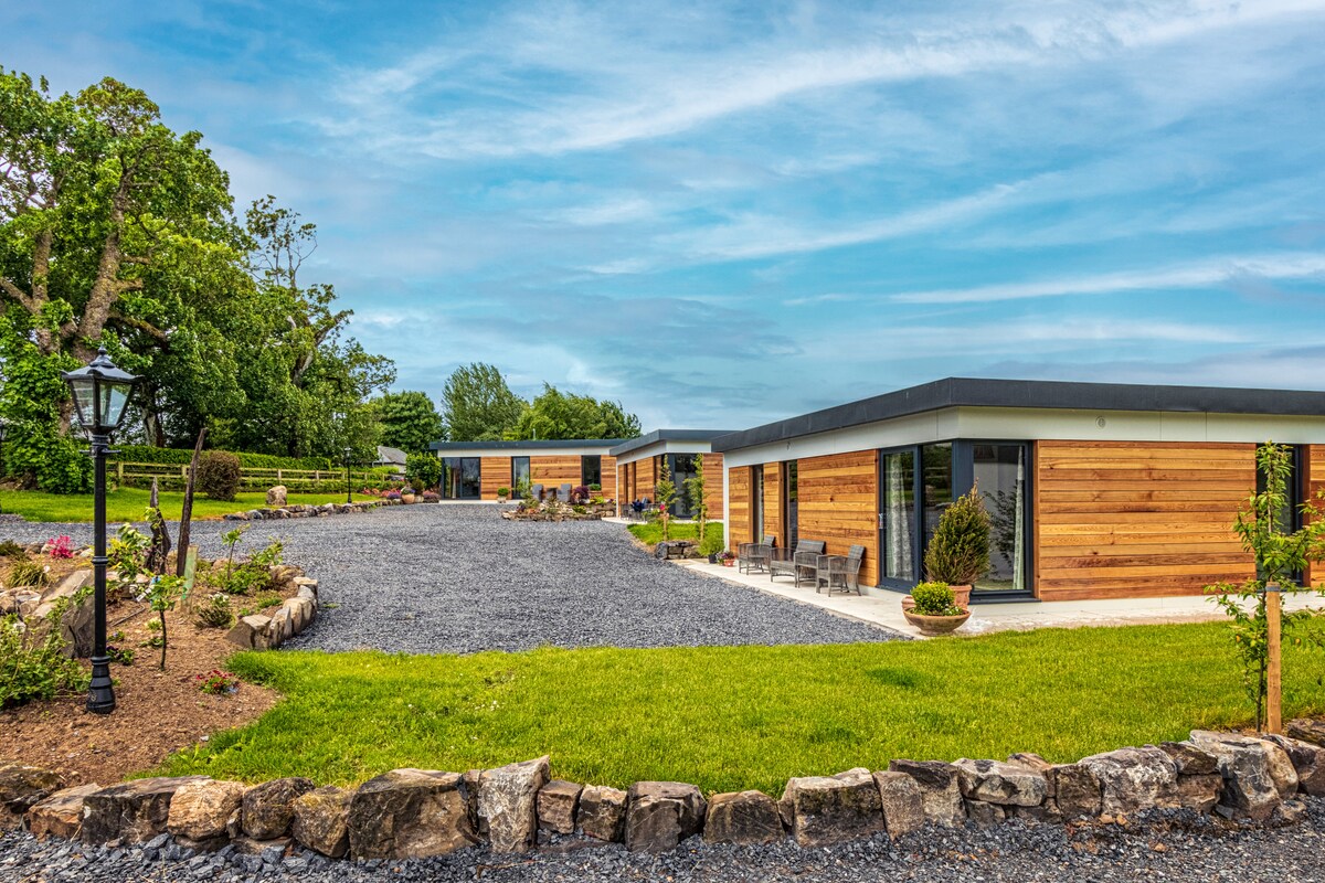 The exterior of the lodge is depicted, showcasing wooden-clad structures arranged around a gravel driveway. Lush gardens are visible alongside, featuring various plants and landscaping elements. The sky is overcast, providing a tranquil backdrop to the accommodation.
