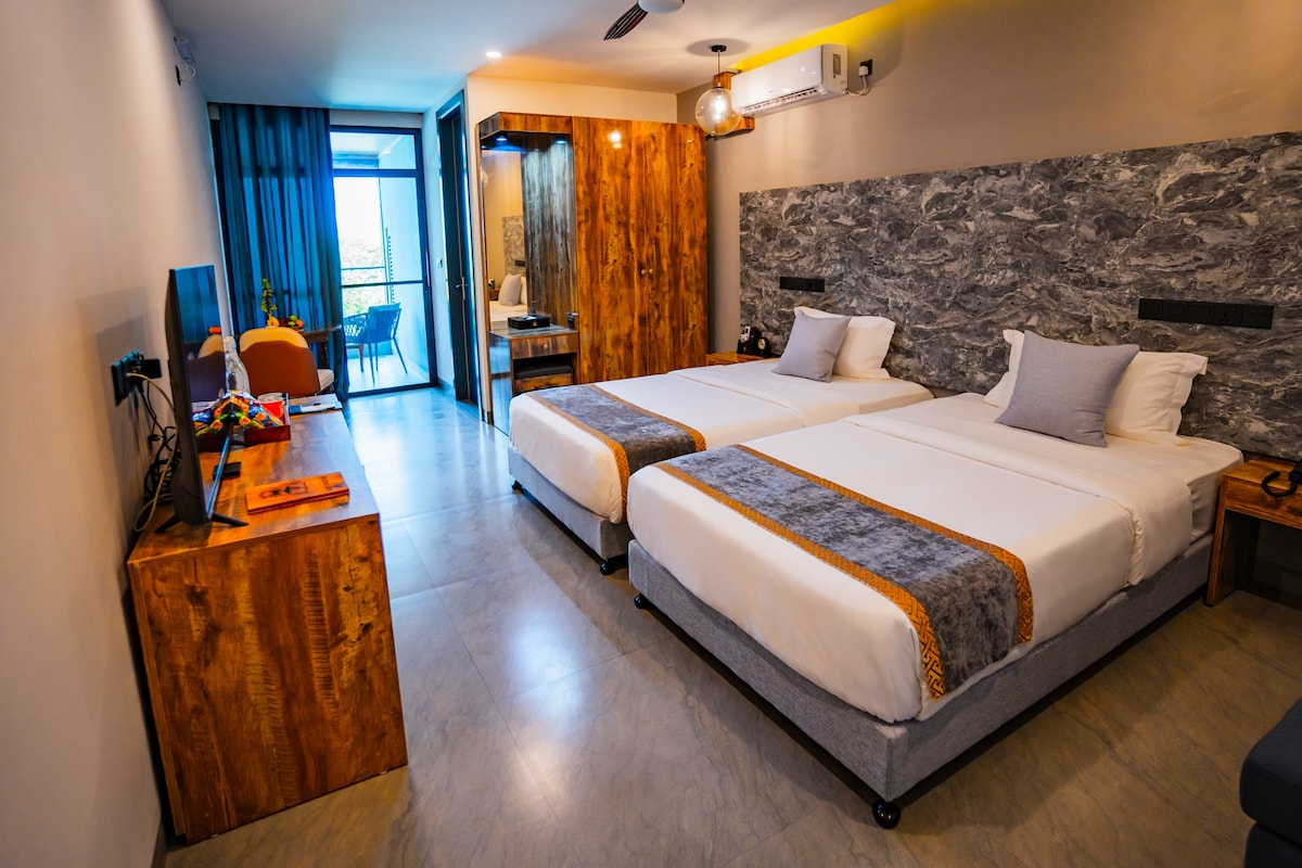 A well-appointed room features two spacious beds with light linens. Natural wood accents are included in the furniture, complemented by a modern television. Large sliding doors lead to a private balcony, allowing natural light to enhance the open layout.