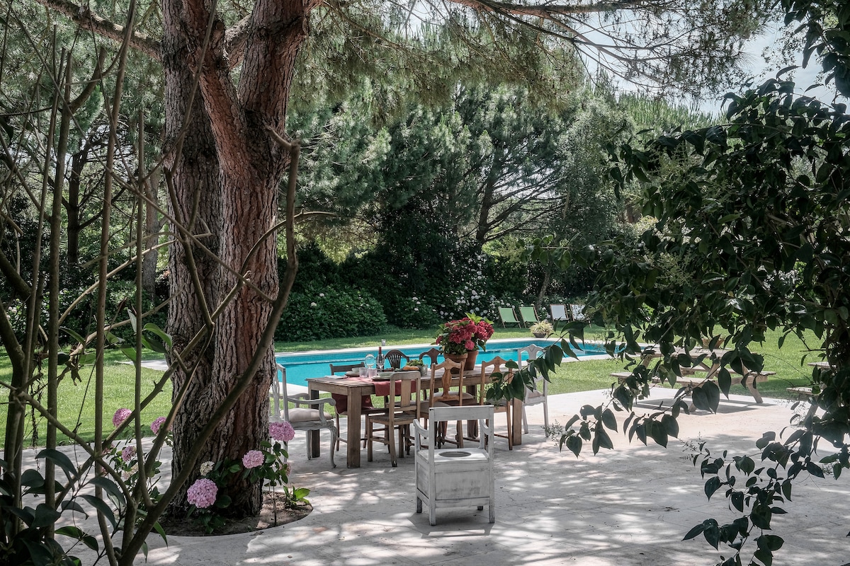 A shaded outdoor dining area is set with a wooden table surrounded by chairs, offering a peaceful view of the pool and lush greenery. Colorful flowers and large trees add to the natural setting, creating a serene atmosphere for gatherings.