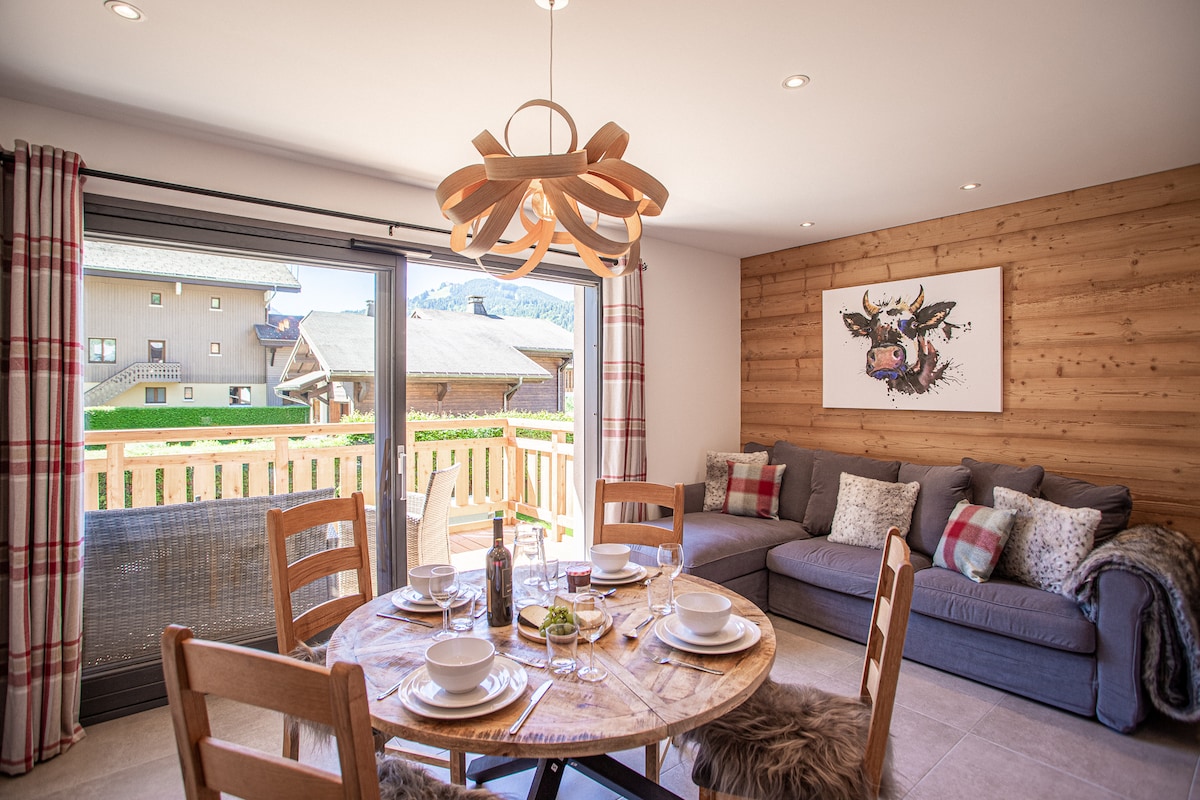 An open-plan dining area features a round wooden table set for four, complemented by a large L-shaped sofa adorned with cushions. Light enters through sliding glass doors, leading to a private balcony. A contemporary light fixture hangs above the table, creating a cozy ambiance.