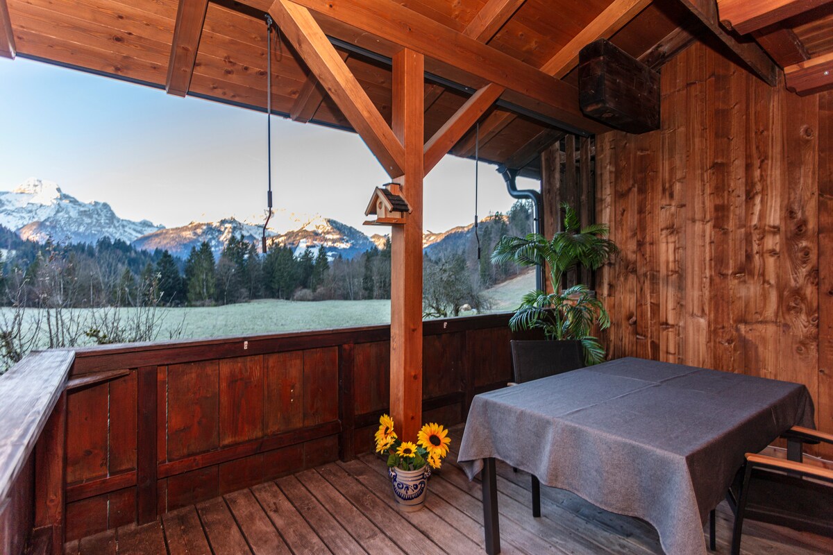 A wooden terrace features a table set with a gray cloth and a pot of sunflowers. Surrounding mountains are visible in the background, complemented by a clear sky. A potted plant adds a touch of greenery, and the rustic wooden walls enhance the natural ambiance.